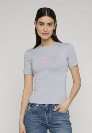 Donna con capelli scuri lisci che indossa una maglietta aderente a maniche corte grigio chiaro con logo rosa e jeans blu, posando con una mano in tasca.