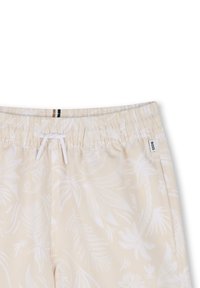Shorts de bain beiges avec un motif de feuilles tropicales blanches, taille élastique, fermeture par cordon de serrage, et une petite étiquette avec le logo de la marque.
