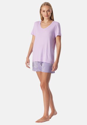 Femme debout pieds nus portant un haut violet clair à manches courtes en V et un short assorti à motifs sur un fond uni.
