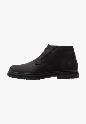 Bota de tobillo de cuero negro con cordones en la parte delantera, suela gruesa y logo del árbol Timberland estampado en el lado del talón.