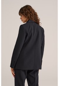 Kvinna med lockigt brunt hår som bär en figurnära svart kavaj och matchande byxor, stående mot en enfärgad beige bakgrund, vänd åt vänster.