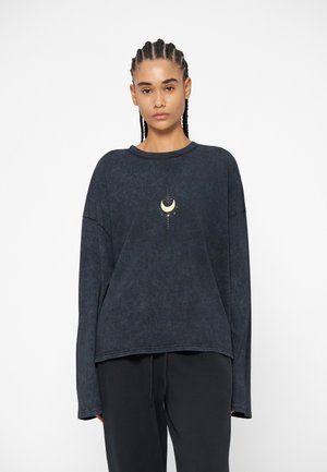 Femme aux cheveux tressés portant un haut ample à manches longues sombre avec un petit motif de croissant de lune et d'étoile au centre.