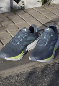 Scarpe da corsa blu con tomaia in mesh, accenti verdi e suola bianca. Presentano tre strisce scure. Design leggero, superficie texture per una migliore aderenza.