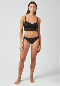Zwarte kant bralette met golvende randen en dunne banden, gecombineerd met bijpassende kanten slipjes. Geweven bloemmotief overal.