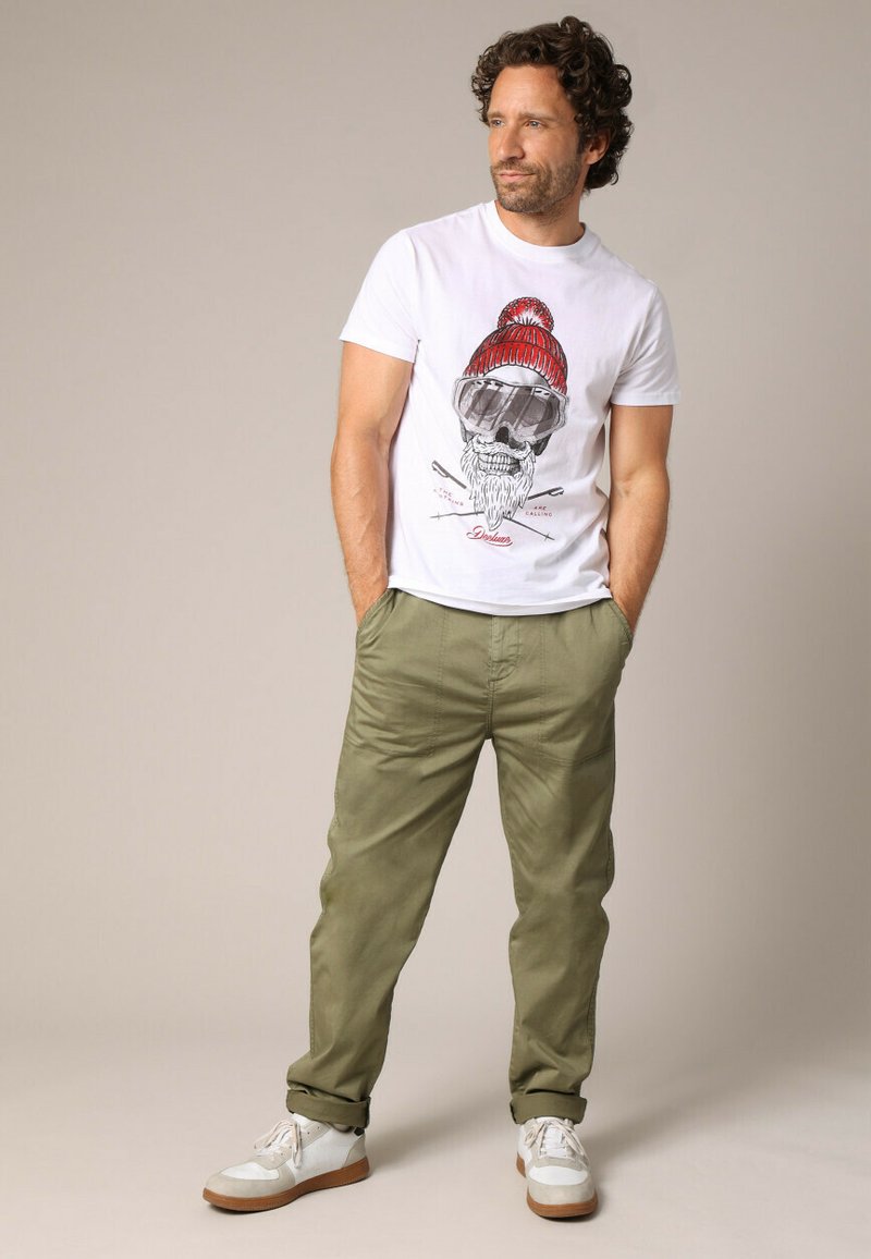 T-shirt blanc avec un motif de crâne portant un bonnet rouge et des lunettes; associé à un pantalon tapered vert olive et des baskets blanches avec des accents beiges.