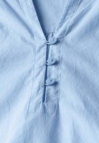 Camicia azzurra chiara con colletto a V, tre bottoni, tessuto testurizzato e una piccola fessura sotto i bottoni.