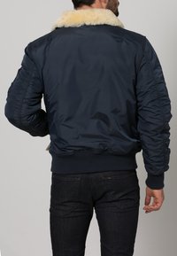 Marinblå bomberjacka med en krage av fuskpäls, ribbade ärmslut och midja. Slät, glänsande tyg med rynkade detaljer på axlarna.