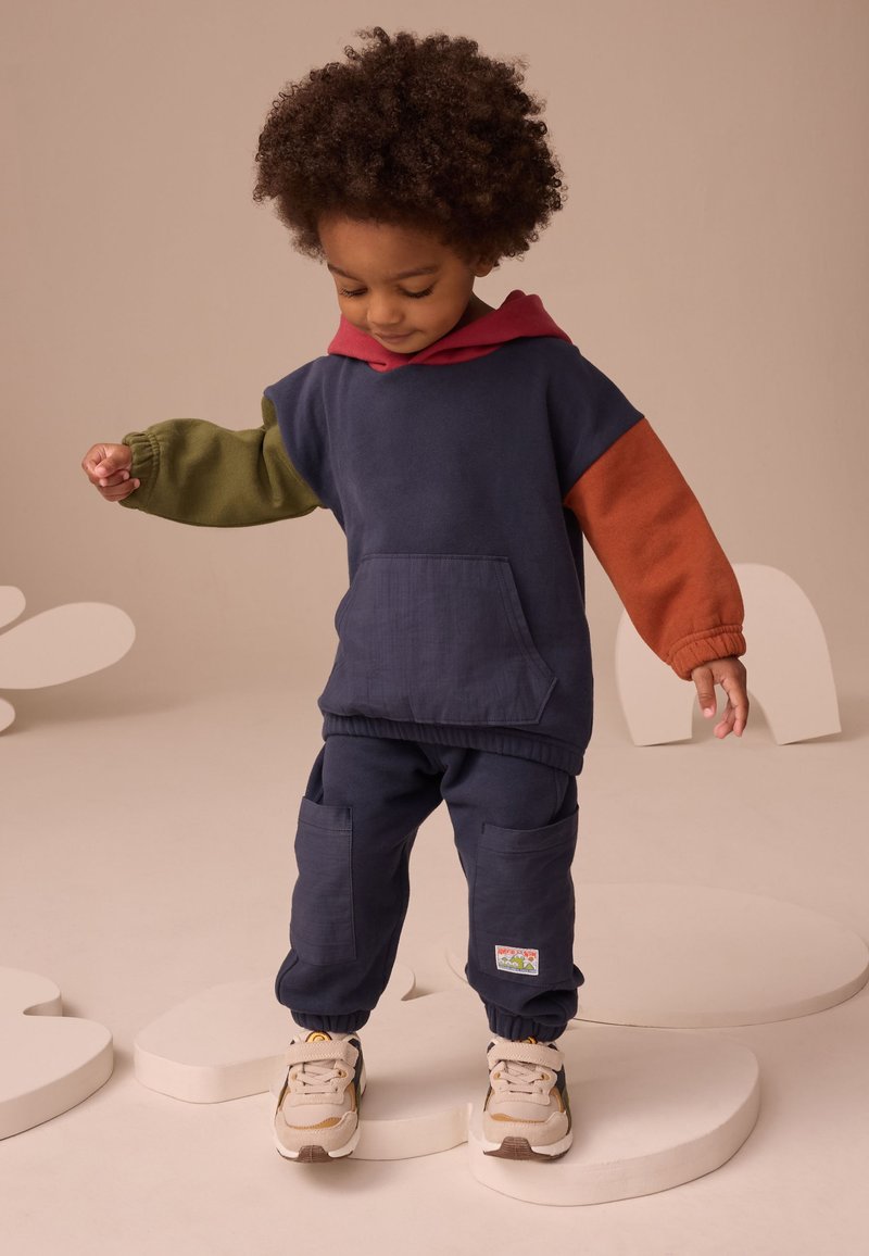 Małe dziecko o kręconych włosach, ubrane w granatową bluzę z kapturem z zielonymi i pomarańczowymi rękawami oraz pasujące spodnie, stojące na białych abstrakcyjnych kształtach.