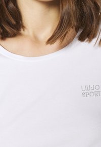 Vrouw met schouderlang bruin haar die een wit shirt draagt met strass-tekst "Liu Jo Sport" op de borst.