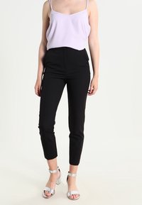 Kvinna klädd i en lila ärmlös topp, svarta slim-fit cropped byxor och silverfärgade högklackade sandaler som står mot en vit bakgrund.