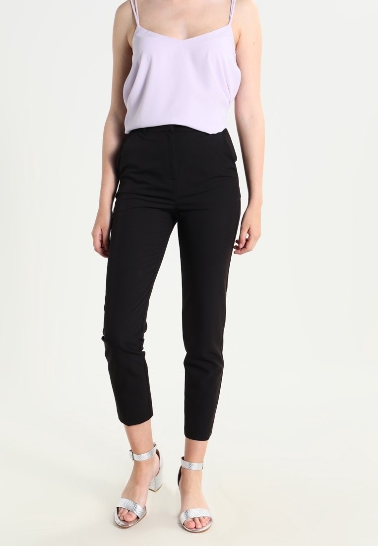 Kvinna klädd i en lila ärmlös topp, svarta slim-fit cropped byxor och silverfärgade högklackade sandaler som står mot en vit bakgrund.