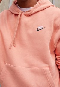 Persikofärgad Nike-hoodie med framficka, dragsko i huvan och broderad logga på bröstet, tillverkad av mjuk bomullsblandning.