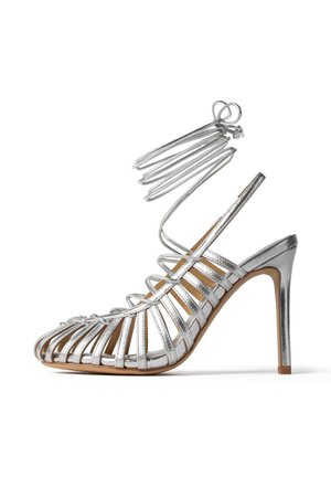 Srebrne sandały na wysokim obcasie z otwartym noskiem, z cienkimi paskami i zapięciem wokół kostki. Szpilka o gładkim wykończeniu i jasnobeżowa wkładka.