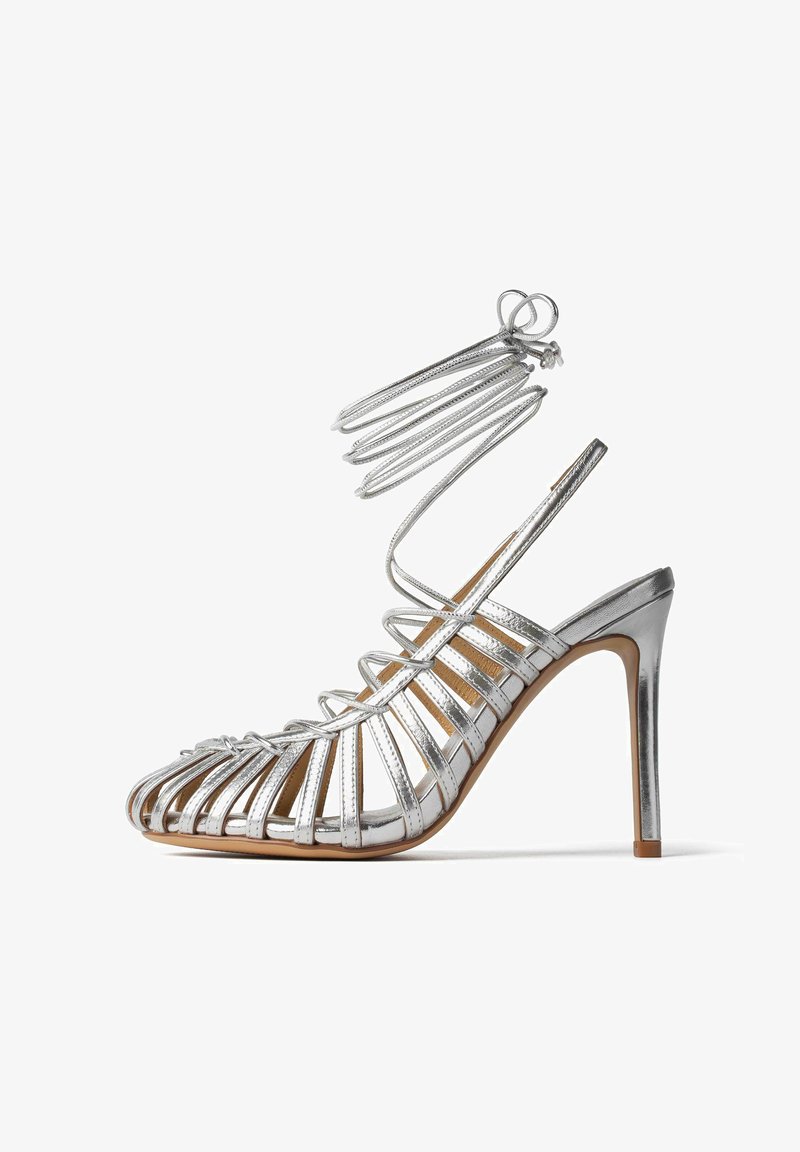 Sandalo argentato con design open-toe, caratterizzato da sottili cinturini e una fascia alla caviglia. Tacco a stiletto con finitura liscia e soletta beige chiaro.