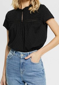 Blouse noire à manches courtes avec un col à volants, des points texturés et une patte de boutonnage frontale. Associée à un jean bleu clair taille haute.