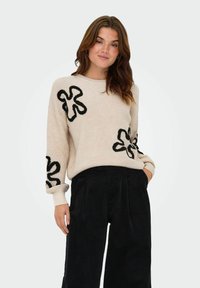 Beige knit sweater with black floral embroidery on sleeves, crew neck, and relaxed fit. Paired with black wide-leg corduroy pants.