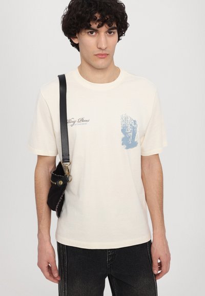 T-shirt en coton crème avec un imprimé graphique bleu, à manches courtes et col rond. Associé à un jean en denim foncé et un sac bandoulière noir.
