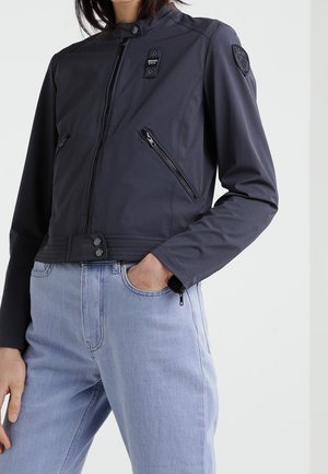 Personne portant une veste noire ajustée zippée avec deux poches diagonales zippées et un jean bleu clair, main dans la poche.