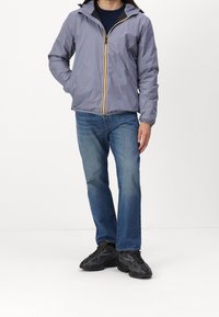 Veste gris clair zippée avec capuche. Présente un accent jaune le long de la fermeture éclair. Portée avec un jean bleu et des chaussures de sport noires. Texture lisse.