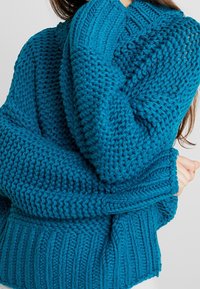 Pull en maille turquoise avec une texture épaisse, des poignets côtelés et une coupe décontractée. Remarquable pour son fil épais et sa construction robuste.