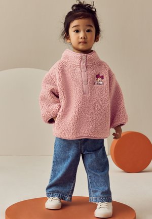 Kleinkind trägt einen pinken Fleecepullover mit Minnie-Maus-Stickerei, blaue Jeans und weiße Turnschuhe, steht auf einer orangefarbenen runden Plattform.