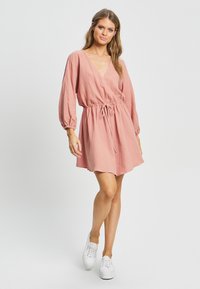 Roza obleka s teksturo in dolgimi rokavi, z vezanjem v pasu in raztegnjenim krilom. V kombinaciji z belimi supergami. Preprost in sproščen dizajn.