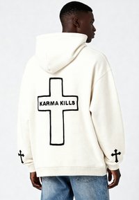 Felpa con cappuccio crema, caratterizzata da una grande croce nera e la scritta "KARMA KILLS" sul retro, con croci più piccole su ogni manica.