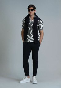 Kurzärmliges graues Polohemd mit weißen abstrakten Mustern, marinefarbener ärmelloser Pullover, dunkle Hose und weiße Sneakers; minimalistische und moderne Optik.