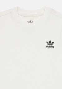 Λευκό βαμβακερό T-shirt με στρογγυλή λαιμόκοψη και μικρό μαύρο λογότυπο Adidas με τρία πέταλα πάνω από το όνομα της μάρκας στο αριστερό στήθος.