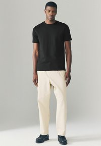 Sort bomulds T-shirt med korte ærmer, parret med beige løse bukser og sorte sko, stående mod en lys grå baggrund.