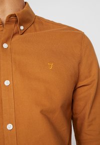 Bruine button-up shirt van katoen met een zachte textuur. Heeft een button-down kraag en een subtiele gele geborduurde logo op de borst.