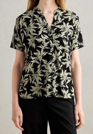 Vrouw draagt een zwart T-shirt met korte mouwen met een lichtgroen tropisch bladerenpatroon en zwarte broek, tegen een effen achtergrond.