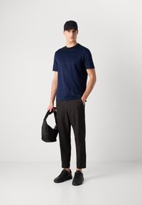 Jovem usando uma t-shirt às riscas azul-escura, calças pretas à altura dos tornozelos, ténis pretos e um boné preto, segurando uma mala preta em um ambiente de estúdio neutro.