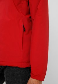 Red waterproof jacket with a smooth texture, elastic cuffs, and adjustable drawstring at the hem. Features a side pocket for storage.