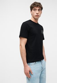 Νέος άνδρας με μαύρο μπλουζάκι με μικρό λογότυπο στο στήθος, ανοιχτόχρωμο μπλε τζιν, στέκεται με το ένα χέρι στην τσέπη, κοιτάζοντας πλάγια.