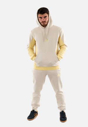 Crème en gele hoodie met kangoeroezak, bijpassende joggers en zwarte sneakers met bruine accenten. Bevat adidas-logo's op de kleding.