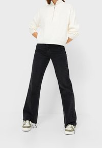 Witte vest met rits, zwarte uitlopende jeans en groen-witte sneakers. De outfit heeft een ontspannen pasvorm en een minimalistisch ontwerp.