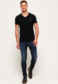 Camiseta negra de cuello en V, de mangas cortas, material de algodón, diseño ajustado, combinada con jeans oscuros y botas marrones hasta el tobillo, de pie contra un fondo liso.