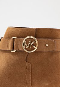 Bruine suède laars met een gouden ronde gesp waarop de initialen "MK" staan, aan een bandje nabij de bovenrand.