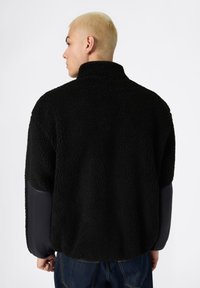 Chaqueta de forro polar negra con cuello alto, cuerpo texturizado y mangas lisas negras en contraste. Diseño casual con un ajuste holgado.