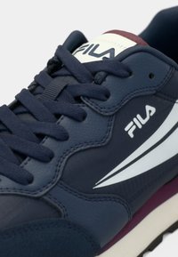 Navy blue athletic shoe with textured synthetic leather upper, white accents, dark laces, and raised branding. Rubber sole with multi-pattern tread.