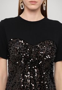 Femme portant une chemise noire avec un grand motif noir à sequins sur la poitrine et de longues boucles d’oreilles en chaînes dorées et argentées