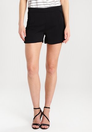 Femme portant un short noir taille haute, des sandales noires à talons avec des lanières, et un haut blanc et noir rayé rentré, sur fond blanc.