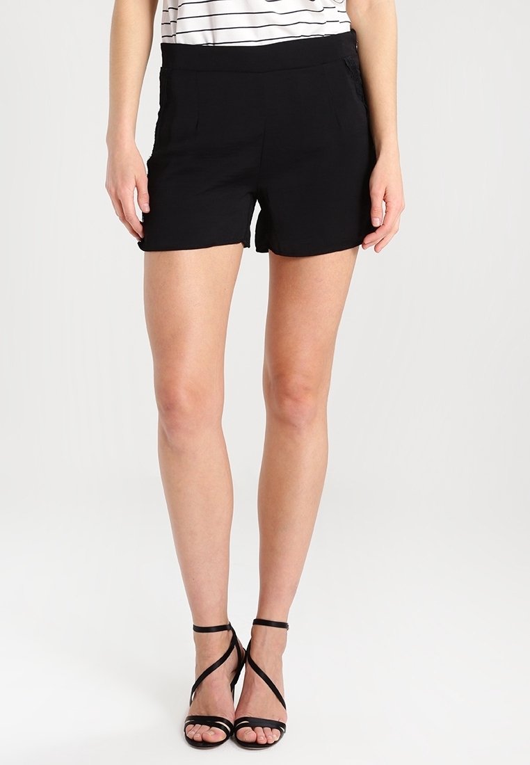 Femme portant un short noir taille haute, des sandales noires à talons avec des lanières, et un haut blanc et noir rayé rentré, sur fond blanc.