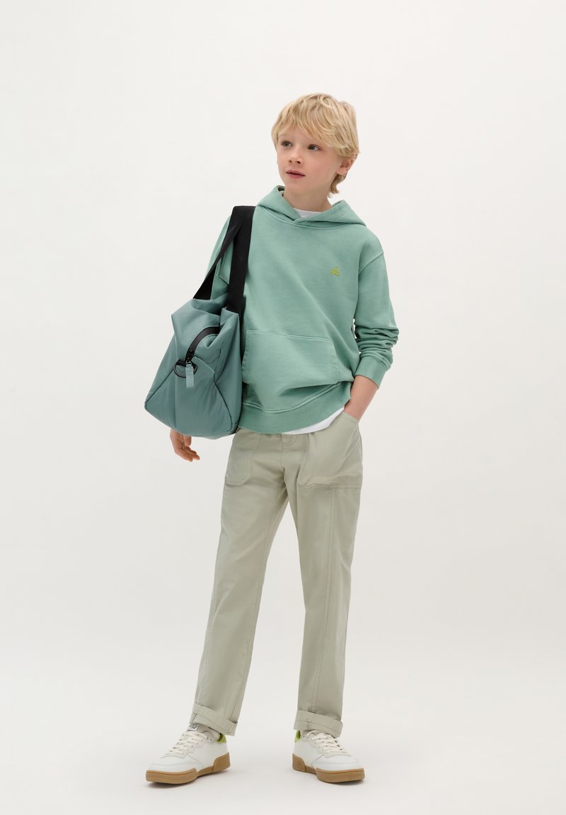 Felpa verde menta con tasca a marsupio, abbinata a pantaloni beige chiaro. La modella porta una borsa turchese con una tracolla nera e una zip. Sneakers bianche.