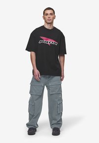 T-shirt oversized preto com logotipo branco e vermelho e calças cargo cinzentas com vários bolsos. Usado com ténis escuros num fundo liso.