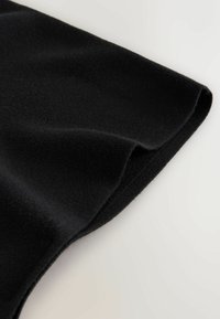 Svart tyg, mjuk textur, med en slät, något böjd kant och prydlig sömnadsdetalj. Nära vy av plagget.