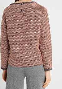 Sweter dzianinowy w fakturowanym koralu z czarnym wykończeniem przy dekolcie i zapięciem na guziki z tyłu. W zestawie z spodniami w jodełkę.