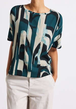 Personne portant un haut à manches courtes à motif abstrait bleu canard et crème avec un pantalon beige clair, les mains dans les poches, sur fond uni.