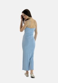 Robe maxi ajustée de couleur bleu clair avec des fines bretelles, texture lisse et design au milieu du dos. Accompagnée de chaussures béiges à enfiler.